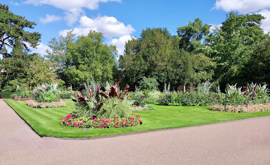 A well-maintained garden scene featuring vibrant flower beds with pink and white blooms arranged around lush, trimmed grass lawns. The garden includes neatly edged flower beds with a variety of plants