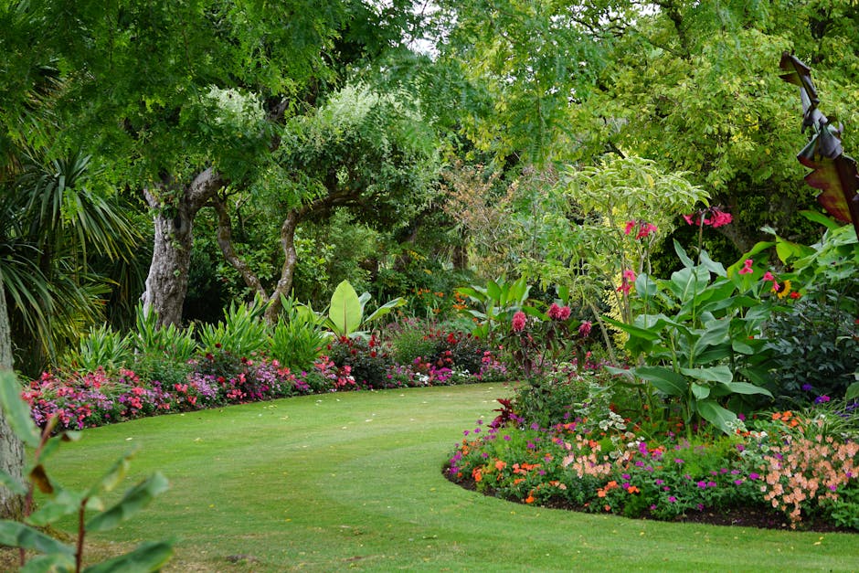 A well-maintained garden scene featuring vibrant flower beds with pink and white blooms arranged around lush, trimmed grass lawns. The garden includes neatly edged flower beds with a variety of plants